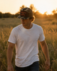 A man walking through a grassy field at sunset, wearing a white crew-neck t-shirt and a dark camouflage trucker hat with a red and black 'CRUSH THE' graphic patch.