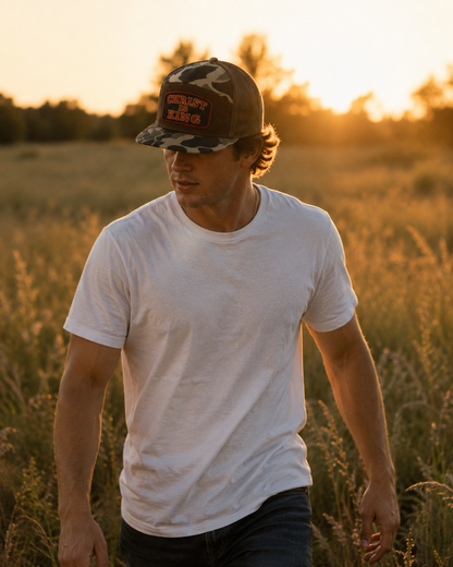 A man walking through a grassy field at sunset, wearing a white crew-neck t-shirt and a dark camouflage trucker hat with a red and black 'CRUSH THE' graphic patch.