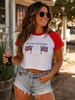 A woman wearing a white and red raglan crop top with the text 'RED WHITE AND BOBBIES' and two American flag graphics, paired with denim shorts and holding a Coca-Cola bottle in an outdoor setting.