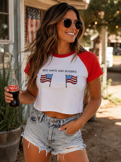 A woman wearing a white and red raglan crop top with the text 'RED WHITE AND BOBBIES' and two American flag graphics, paired with denim shorts and holding a Coca-Cola bottle in an outdoor setting.