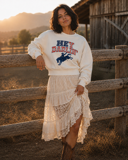 A woman stands outdoors in a rustic barn setting at sunset, wearing a cream sweatshirt with a red, white, and blue western-style graphic and a matching cream lace tiered skirt.