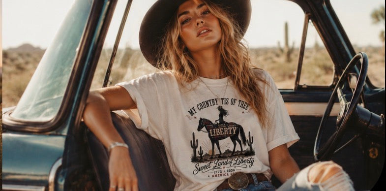 Women's cream vintage Western patriotic shirt with My Country Tis of Thee Liberty cowgirl design worn with cowboy hat sitting in a classic truck in the desert