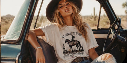 Women's cream vintage Western patriotic shirt with My Country Tis of Thee Liberty cowgirl design worn with cowboy hat sitting in a classic truck in the desert