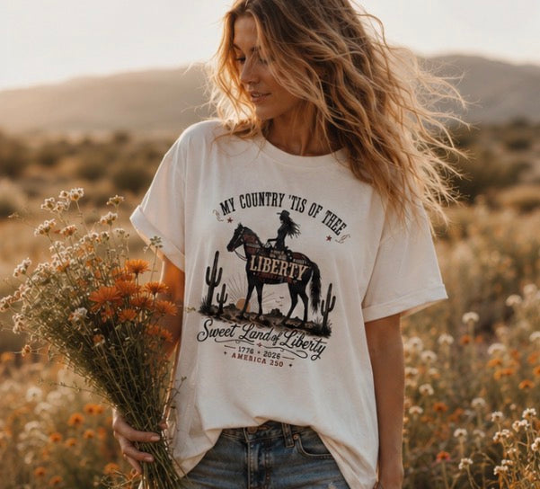 Women's cream relaxed fit patriotic cowgirl tee with Sweet Land of Liberty America 250 graphic worn in a wildflower field with flowing hair