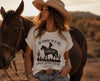 
Women's cream patriotic Western tee with My Country Tis of Thee Sweet Land of Liberty cowgirl graphic worn with cowboy hat and jeans next to a horse at golden hour