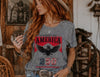 Woman wearing a gray t-shirt with an eagle and 'Celebrating America 250' design in a rustic setting.