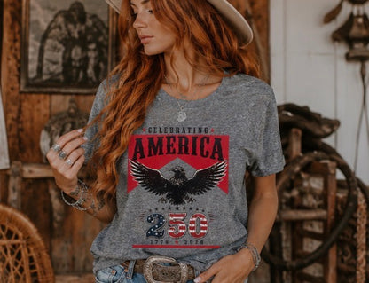 Woman wearing a gray t-shirt with an eagle and 'Celebrating America 250' design in a rustic setting.