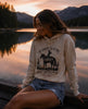 Woman wearing a hoodie with a graphic design sitting by a lake at sunset.