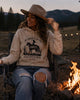 Woman sitting by a campfire wearing a hoodie with a graphic design, surrounded by nature.