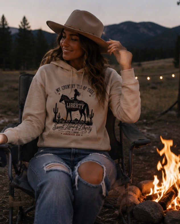 Woman sitting by a campfire wearing a hoodie with a graphic design, surrounded by nature.