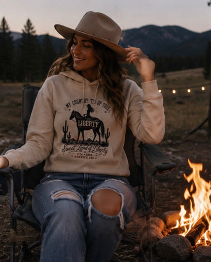 Woman sitting by a campfire wearing a hoodie with a graphic design, surrounded by nature.