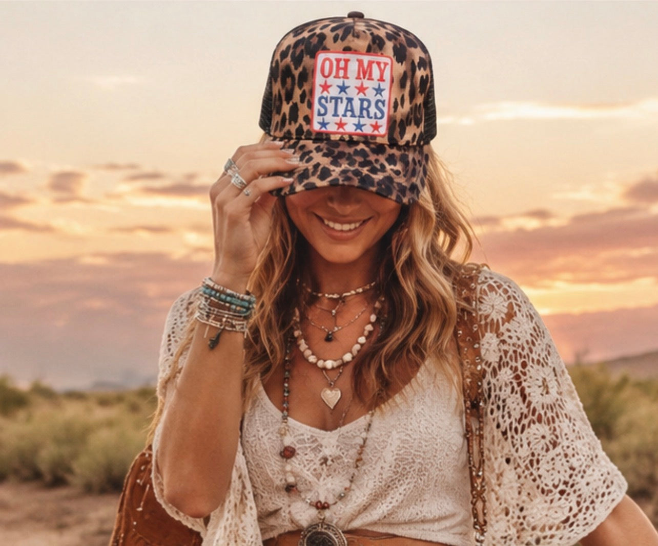 Woman wearing a leopard print cap with 'OH MY STARS' text, sitting outdoors at sunset.