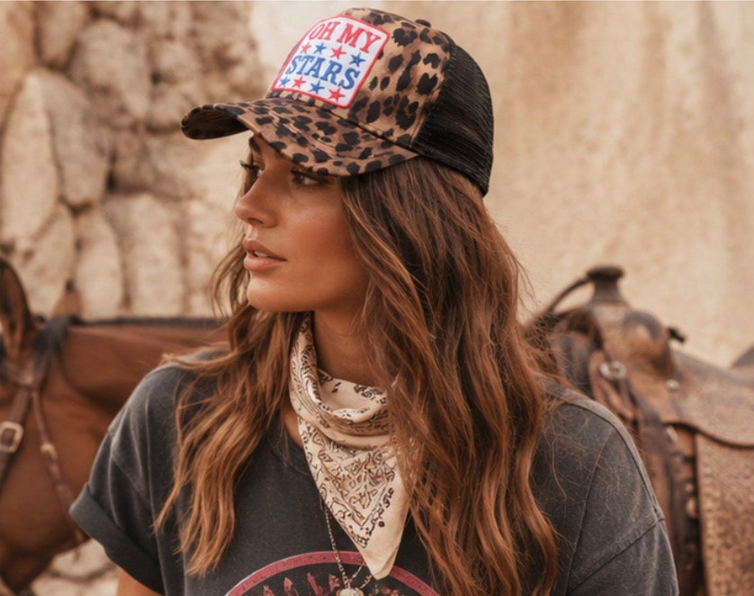 Woman wearing a leopard print cap with a star design and a bandana, standing in front of horseback riding equipment.
