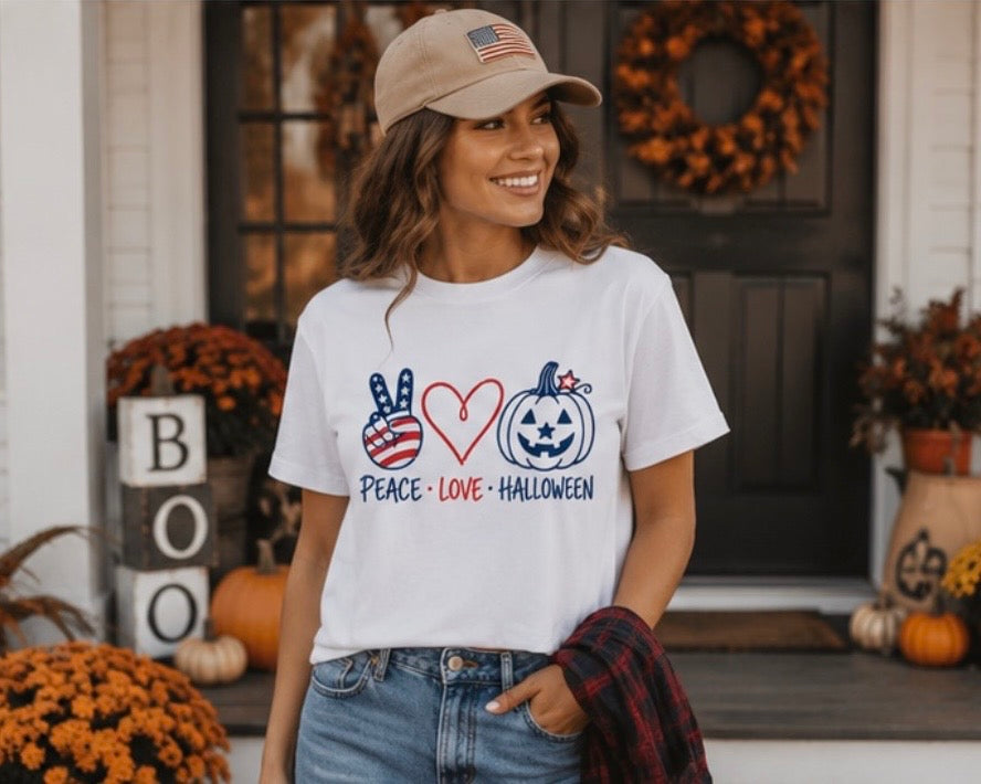 A woman wearing a white graphic t-shirt with a patriotic Halloween design, standing outdoors on a decorated porch. The graphic features a peace sign with an American flag pattern, a red heart, a jack-o'-lantern with stars, and the words 'PEACE • LOVE • HALLOWEEN'.