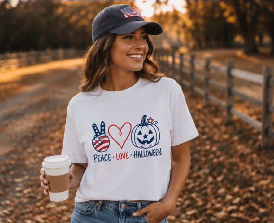 A woman wearing a white graphic t-shirt with a patriotic Halloween design, standing outdoors on a leaf-covered path. The graphic features a peace sign with an American flag pattern, a red heart, a jack-o'-lantern with stars, and the words 'PEACE • LOVE • HALLOWEEN'.