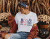 A woman wearing a white graphic t-shirt with a 'Peace Love Halloween' design in red, white, and blue, styled with a rust-colored knit cardigan, jeans, and a patriotic headband in an autumn-themed setting.