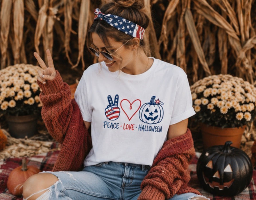 A woman wearing a white graphic t-shirt with a 'Peace Love Halloween' design in red, white, and blue, styled with a rust-colored knit cardigan, jeans, and a patriotic headband in an autumn-themed setting.