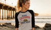 A woman wearing a gray and black raglan t-shirt with a 'MAKE CALIFORNIA GREAT AGAIN' graphic, standing on a rocky beach with a pier in the background.