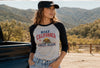 A woman wearing a black baseball cap and a heather gray and black raglan t-shirt with 'MAKE CALIFORNIA GREAT AGAIN' and a grizzly bear graphic, standing outdoors in front of a truck and mountains.