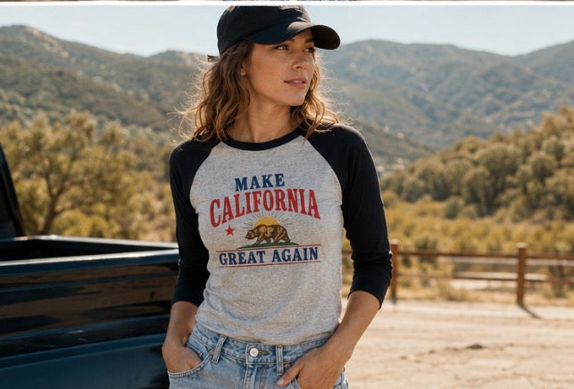 A woman wearing a black baseball cap and a heather gray and black raglan t-shirt with 'MAKE CALIFORNIA GREAT AGAIN' and a grizzly bear graphic, standing outdoors in front of a truck and mountains.