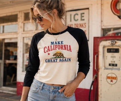 A woman wearing a black and cream raglan baseball t-shirt with a 'Make California Great Again' graphic, standing in front of a vintage gas station and retro gas pump.
