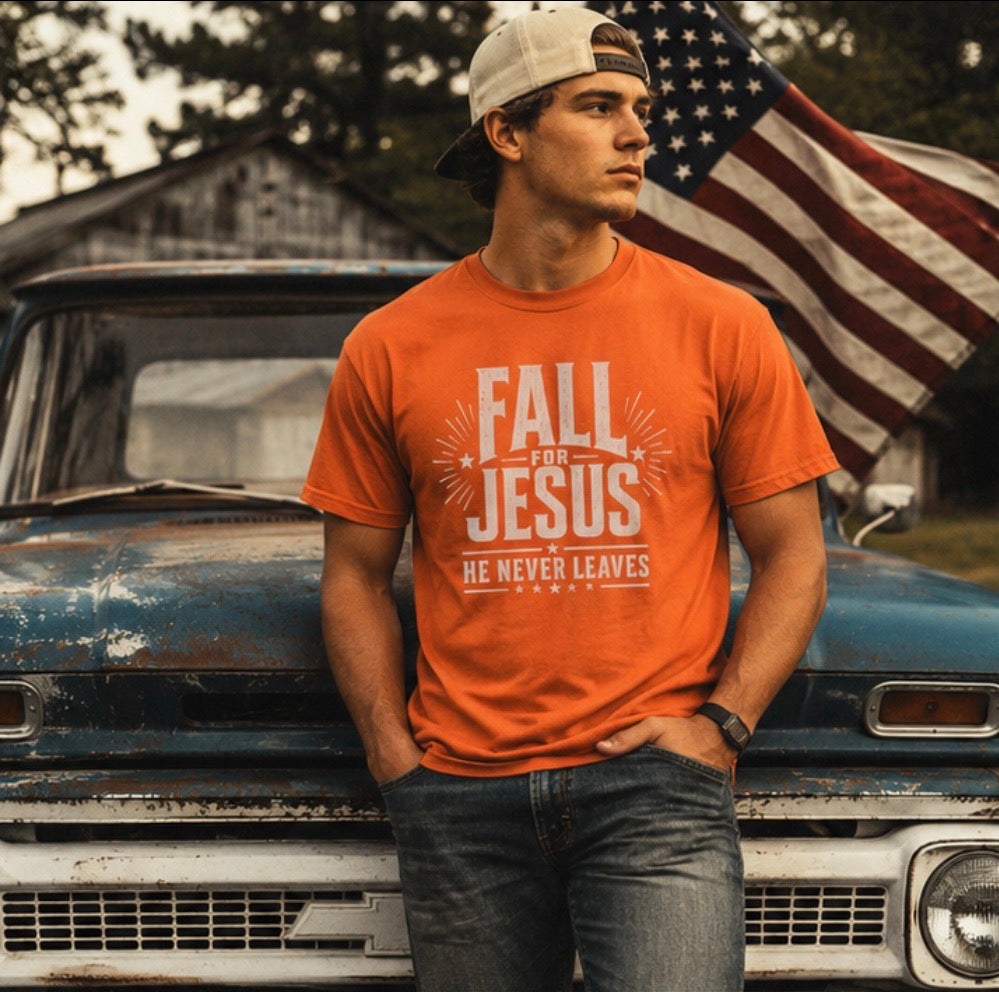 A man wearing a bright orange graphic t-shirt with the text 'FALL OR JESUS HE NEVER LEAVES' stands in front of a vintage blue truck with an American flag in the background.