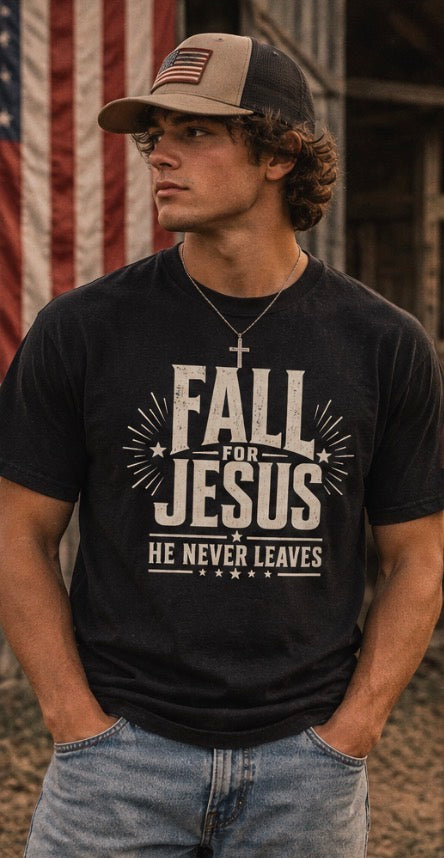 A man wearing a black graphic t-shirt with the text 'FALL FOR JESUS HE NEVER LEAVES' in distressed white lettering, paired with a tan and black baseball cap and light blue jeans, standing in front of an American flag.