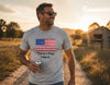 A man wearing a heather gray t-shirt with a printed American flag and the text 'This is a flag, I like it.' walking on a dirt path during sunset.