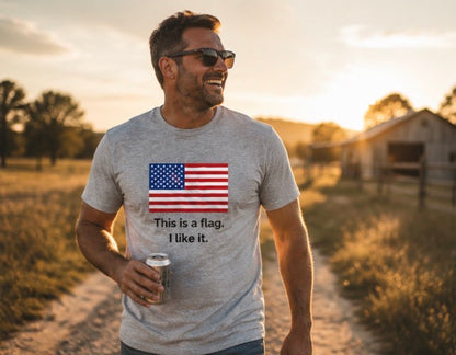 A man wearing a heather gray t-shirt with a printed American flag and the text 'This is a flag, I like it.' walking on a dirt path during sunset.