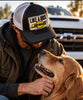 “Man wearing a black and white Like a Rock Jesus trucker hat while petting a golden retriever beside a truck.”
