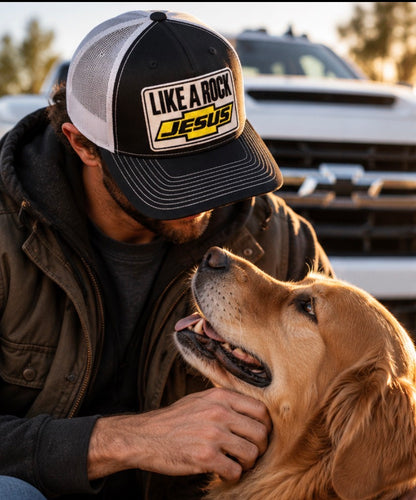 “Man wearing a black and white Like a Rock Jesus trucker hat while petting a golden retriever beside a truck.”
