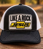 “Front view of Like a Rock Jesus trucker hat with white mesh sides, displayed on a wooden table.”