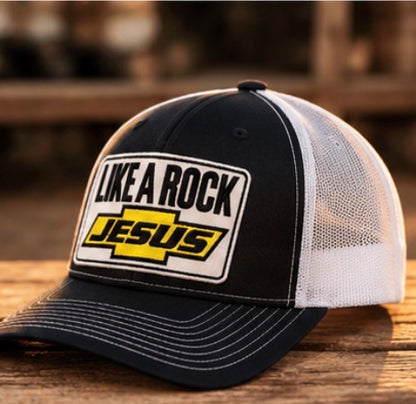 Black and white Like a Rock Jesus trucker hat with yellow patch detail, shown at an angle on a wooden table.”