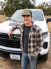 “Man wearing a Like a Rock Jesus camo trucker hat and plaid shirt, leaning against a white pickup truck.”
