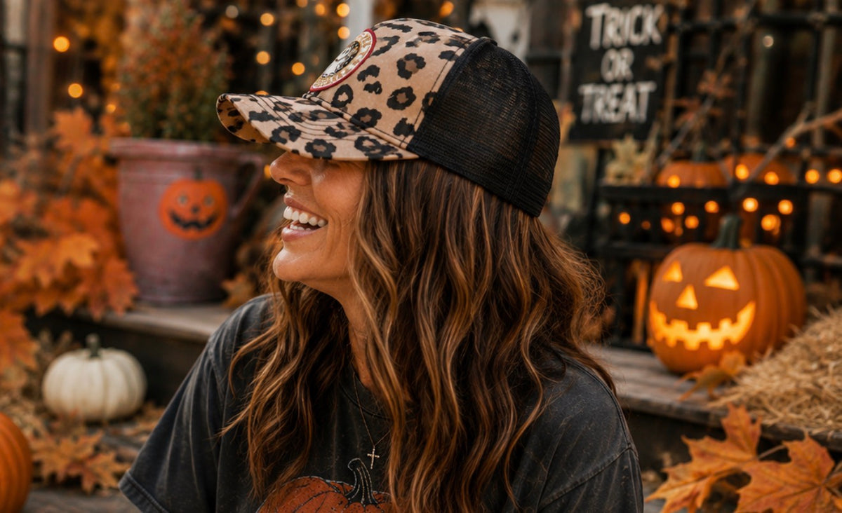 A woman wearing a leopard-print trucker hat with black mesh back panels, smiling in a Halloween-themed outdoor setting with pumpkins and autumn decorations.