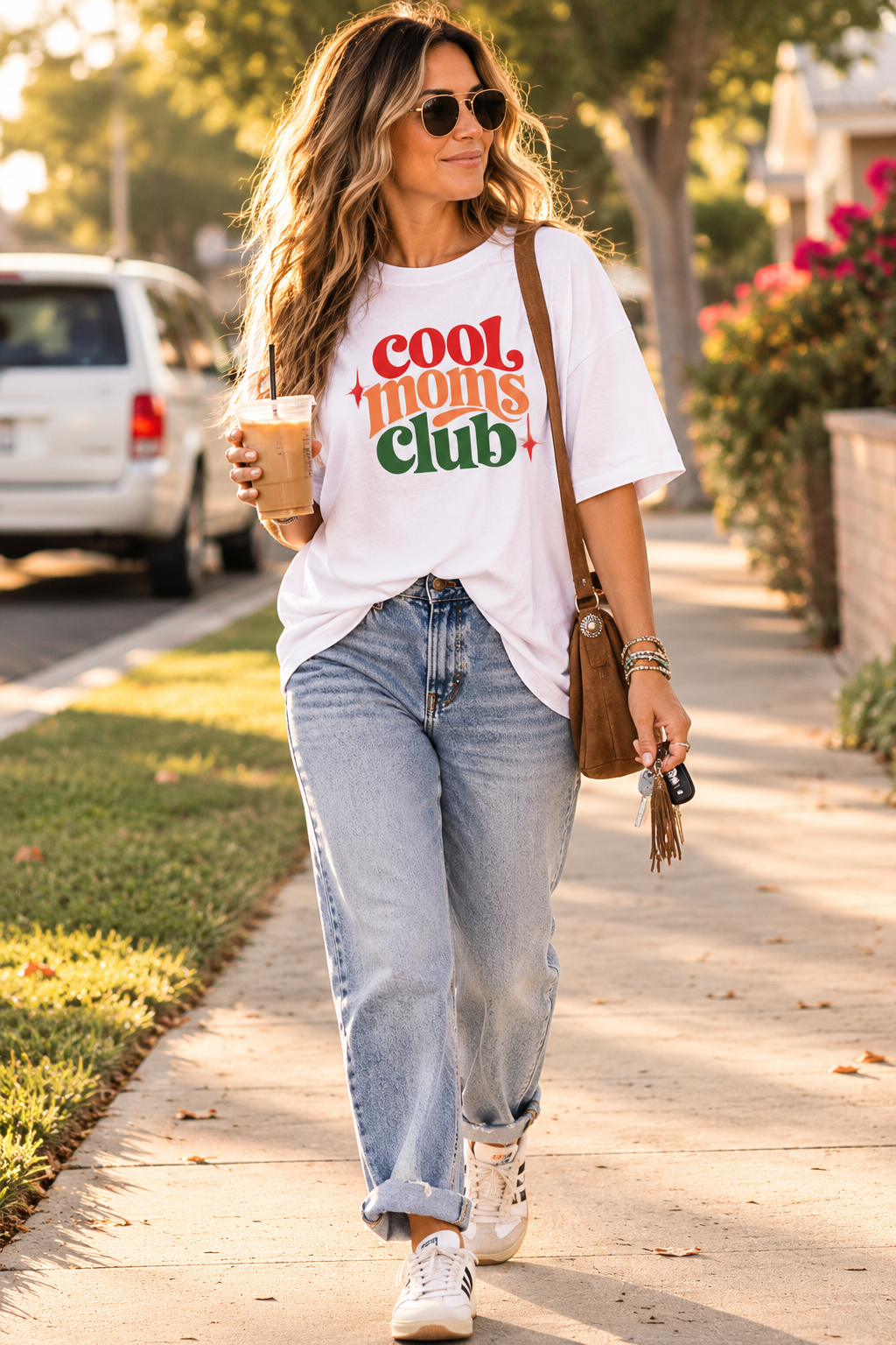 A woman walking on a sunny sidewalk wearing a white graphic t-shirt with 'cool moms club' printed in red, orange, and green, paired with light-wash blue jeans, white sneakers, and a brown shoulder bag.