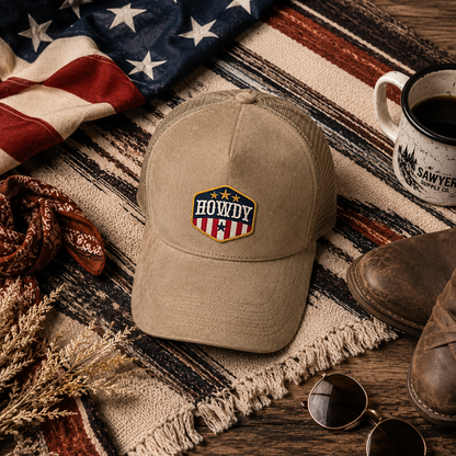 A tan baseball cap with a patriotic embroidered patch reading 'HOWDY' sits on a striped blanket over a wooden surface, surrounded by an American flag, a mug, boots, sunglasses, and dried grass.
