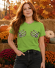 A woman with long red hair stands outdoors in an autumn setting, wearing a bright lime green t-shirt with a graphic of two skeletal hands on the chest, paired with dark gray jeans.
