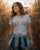 A woman wearing a light gray t-shirt with a graphic of two skeleton hands holding a heart, standing in a cornfield at sunset.
