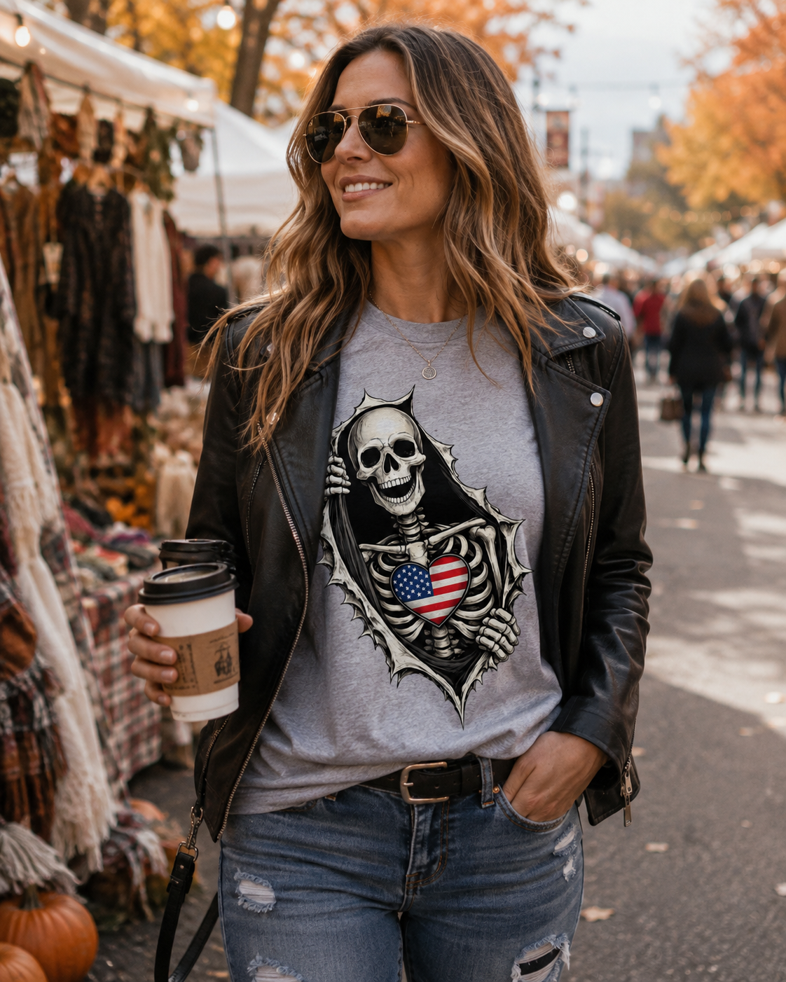 A woman at an outdoor autumn market wearing a black leather jacket, a gray t-shirt with a skeleton and American flag graphic, and blue jeans, holding a coffee cup.