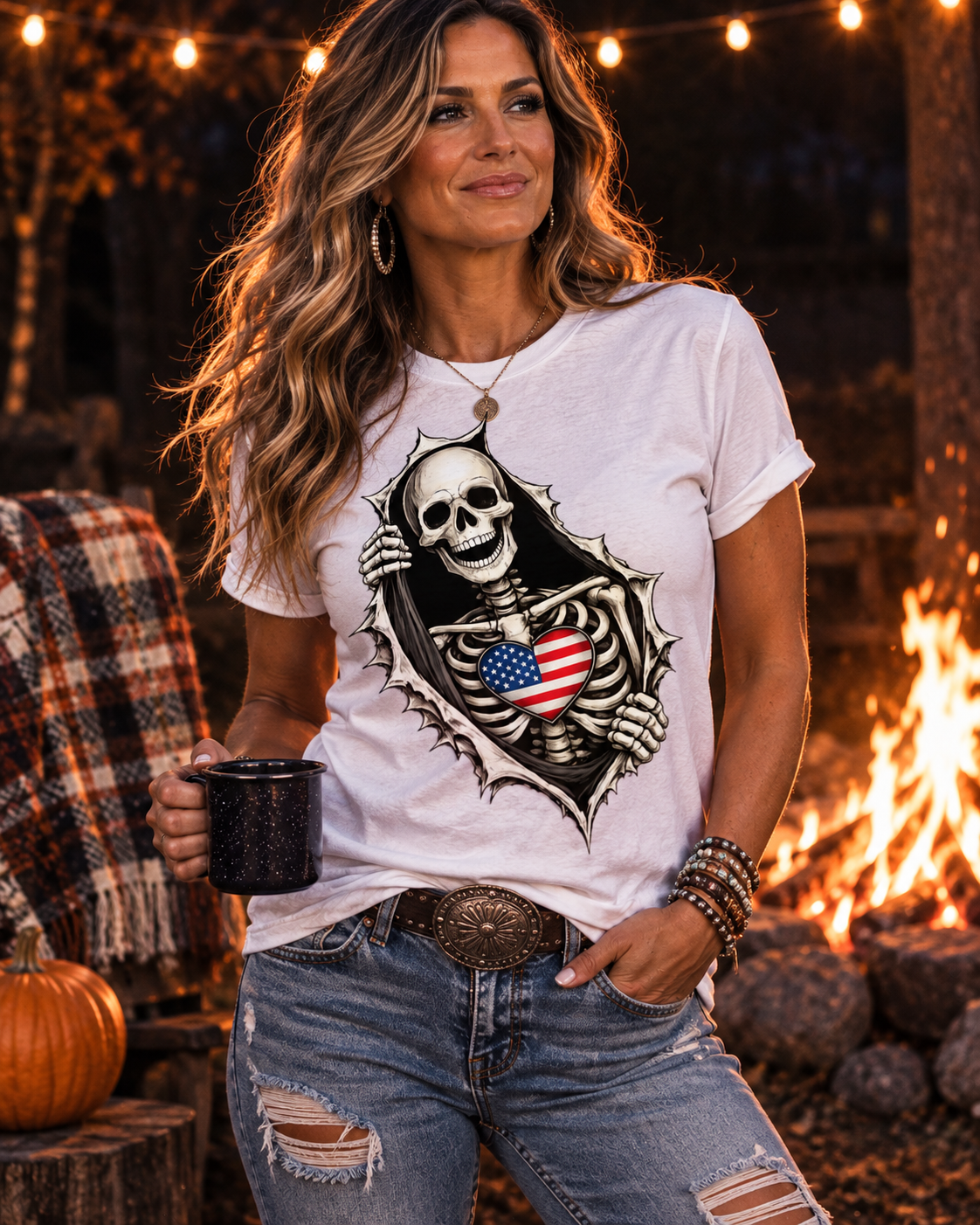 A woman wearing a white graphic t-shirt with a skeleton holding an American flag, paired with distressed blue jeans, standing in front of a warm outdoor campfire at night.