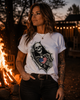 A woman wearing a white graphic t-shirt with a black skeleton and American flag design, standing outdoors near a campfire at dusk.
